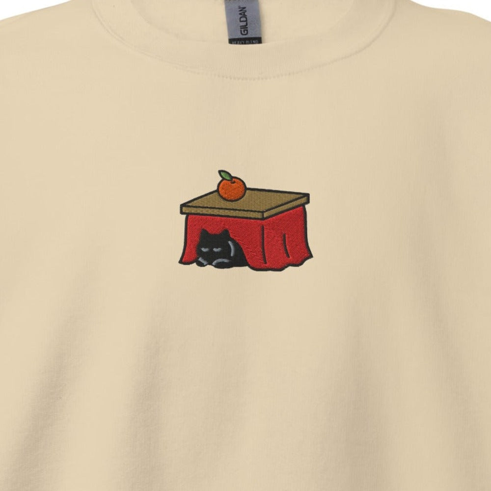 Close-up of the Kotatsu Cat embroidery on a sand crewneck sweatshirt: a black cat sleeping under a red kotatsu with a mandarin orange on top