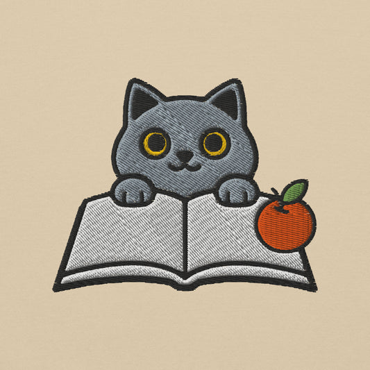 Close-up mockup of the Bookish Cat embroidery on a sand sweatshirt: a grey cat peeking over an open book beside a mandarin orange