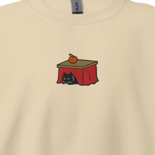 Close-up of the Kotatsu Cat embroidery on a sand crewneck sweatshirt: a black cat sleeping under a red kotatsu with a mandarin orange on top